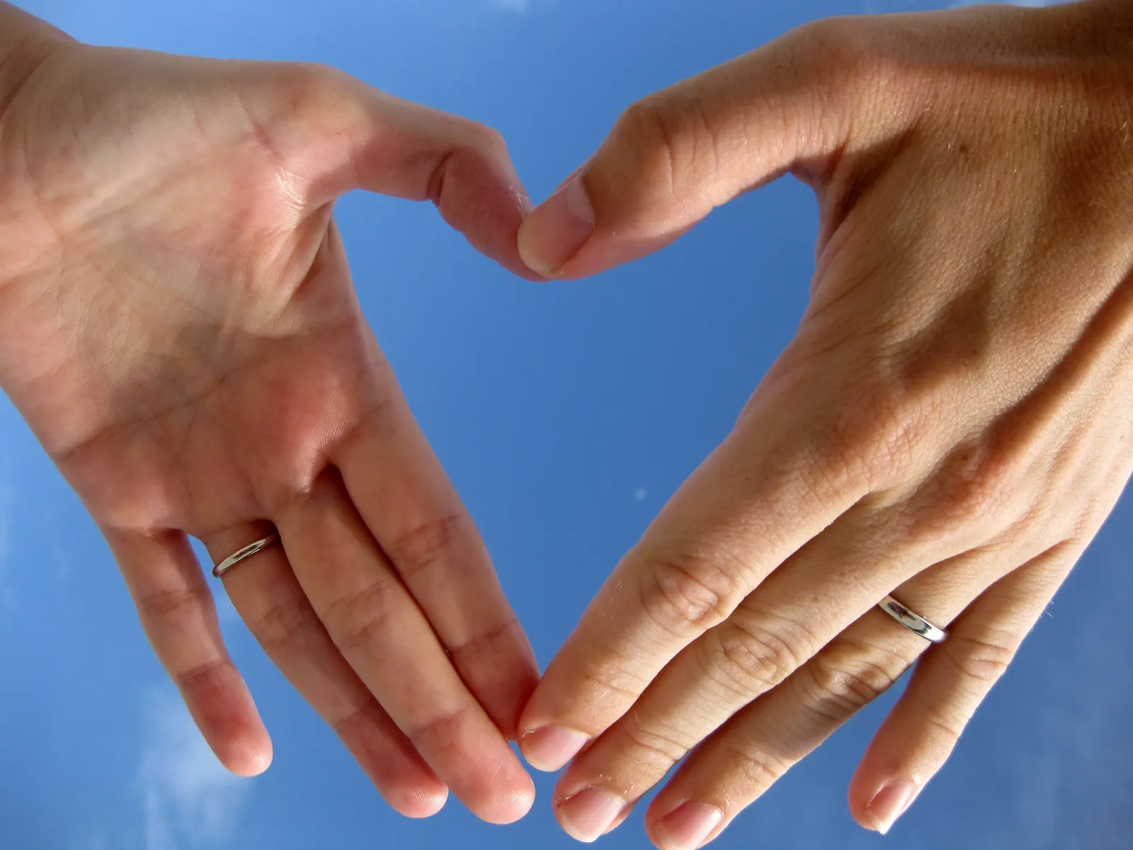 Dos manos con alianzas forman un corazón recortado contra el cielo azul